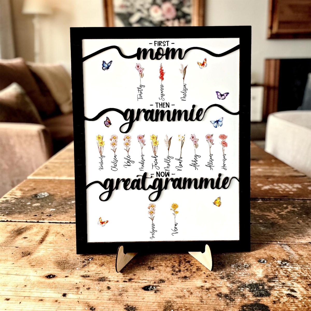 A personalized 'First Mom, Then Grammie, Now Great Grammie' sign displayed on a wooden coffee table in a bright, modern living room. The black-framed 3D artwork features custom family names and birth month flowers, shown in a cozy home environment.