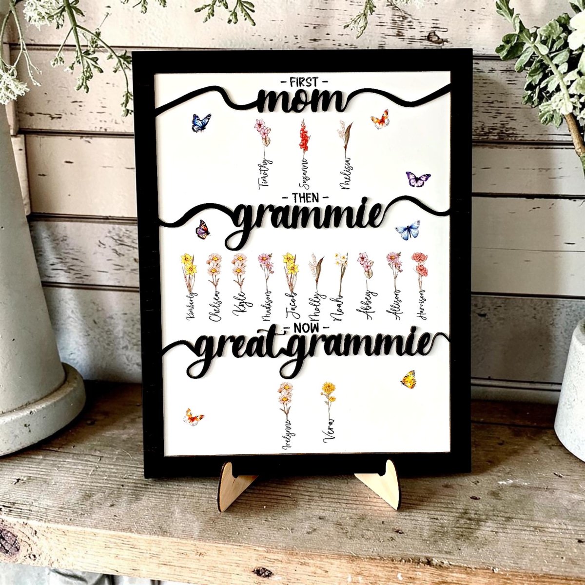 Personalized three-generation family tree sign in a black frame. Featuring 3D black script titles 'First Mom,' 'Then Grammie,' and 'Now Great Grammie' with custom birth month flowers and names. Unique Mother's Day or grandparent gift shown on a wooden display stand.