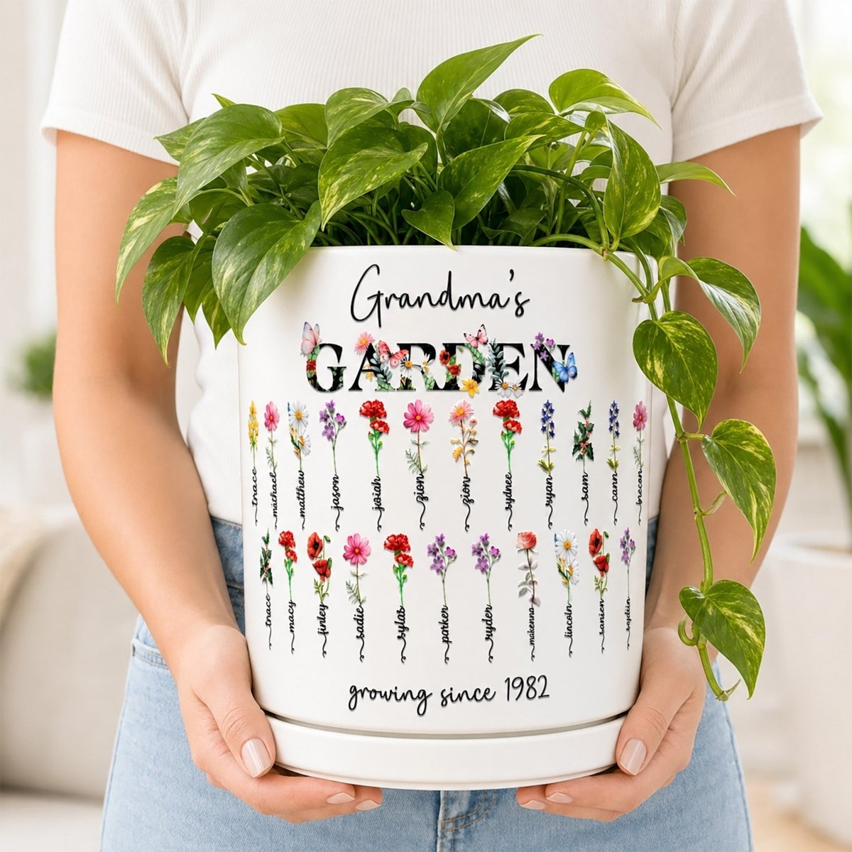 A person holding a large white ceramic planter containing a lush green plant. The custom planter features a "Grandma's Garden" design with two rows of vibrant watercolor birth flowers, personalized family names, and the text "growing since 1982".