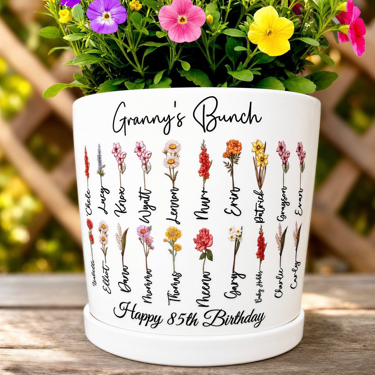 A white ceramic planter pot printed with "Granny's Bunch" and "Happy 85th Birthday" in black script. It features a double-row design of delicate watercolor birth month flowers, each with a personalized name underneath. The pot is filled with vibrant purple, yellow, and pink petunias and sits outdoors against a rustic wooden trellis.