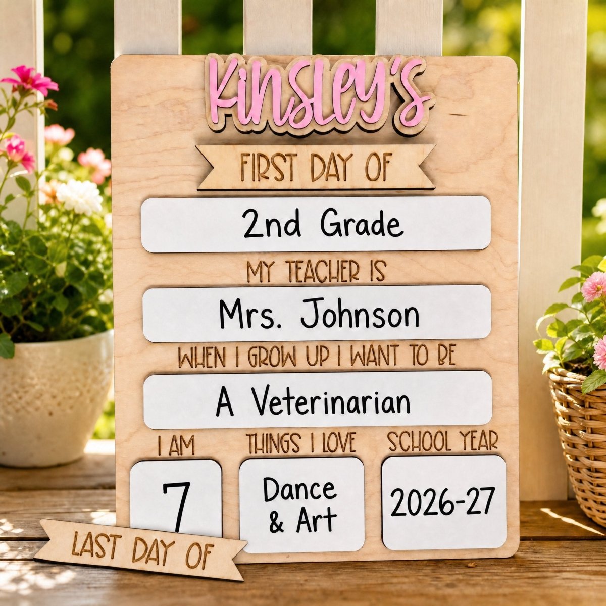 Personalized rectangle first day of school sign displayed outdoors on a wooden surface with lifestyle garden background — maple wood board with pink Kinsley name, filled-in dry-erase sections showing 2nd Grade, Mrs. Johnson, A Veterinarian, and 2026-27 school year